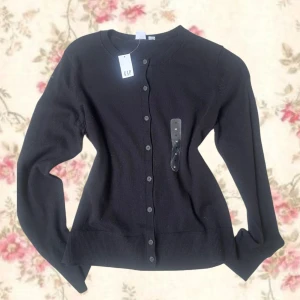 Svart kofta från GAP med knappar - Klassisk svart kofta från GAP med rund halsringning och knäppning hela vägen fram. Långärmad modell i mjukt material, perfekt att slänga över en t-shirt eller klänning. Enkel och stilren design som funkar till det mesta.