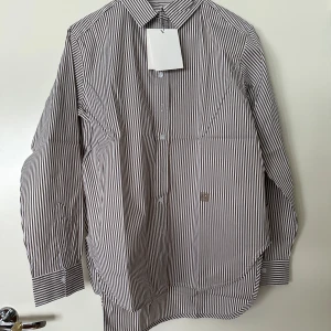 Randig skjorta från Totême i bomull - Snygg skjorta från Totême i vitt och brunt med vertikala ränder. Klassisk krage, långa ärmar och knäppning framtill. Broderad logga på bröstet och tillverkad i mjuk bomull. Perfekt för en clean och stilren look.