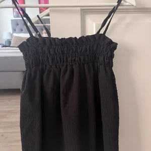 Svart kort topp med smock - Säljer en svart kort topp med smala axelband och smock upptill. Toppen har en lätt textur i tyget och sitter snyggt med en luftig passform. Perfekt för dig som gillar enkel och stilren design.🌺