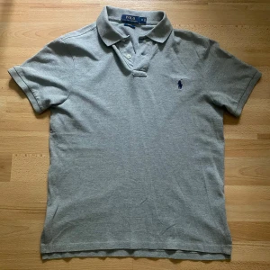 Grå pikétröja från Ralph Lauren - Snygg grå pikétröja från Ralph Lauren med klassisk krage och två knappar framtill. Tröjan har korta ärmar och broderat Polo-logo på bröstet i mörkblått. Knappt använd. Storlek M men sitter mer som S. Hör av dig vid frågor eller fler bilder!