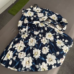 Marinblå blommig långklänning - Säljer en marinblå långklänning med stora vita och ljusgula blommor. Klänningen har långärmade ärmar, hög krage och ett plisserat tyg som ger en snygg siluett. Midjebälte medföljer för extra stilpoäng. Perfekt för dig som vill sticka ut med ett elegant mönster.