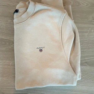 Beige sweatshirt från GANT - Snygg beige sweatshirt från GANT med broderad logga på bröstet. Tröjan har rund halsringning, långa ärmar och ribbade muddar vid ärmslut och nederkant. Tillverkad i mjuk bomull för en skön och avslappnad look.