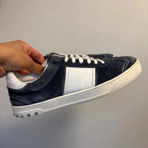 Valentino flycrews mörkblåa/gråa - Snygga Valentino flycrews sneakers i mörkblå/mörkgrå mocka och vitt skinn. Klassisk låg modell med vita snören och coola nitar runt sulan. Skorna har rund tå och platt sula, perfekt för dig som gillar exklusiva detaljer och streetstyle. (skorna saknar endast en nit)