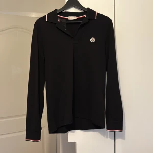 Svart långärmad pikétröja Moncler - Svart långärmad pikétröja från Moncler med klassisk krage och röd-vit-blå rand på krage och ärmslut. Tröjan har Moncler-logga broderad på bröstet och är gjord i mjuk bomull som känns skön mot huden. Perfekt för dig som gillar stilrena och sportiga vibes. Oanvänd