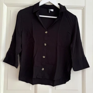 Svart skjorta med trekvartsärm H&M - Svart skjorta från H&M med trekvartsärmar och klassisk krage. Skjortan har knappar framtill och en lite lösare passform. Perfekt till jeans eller kjol för en chill och stilren look.