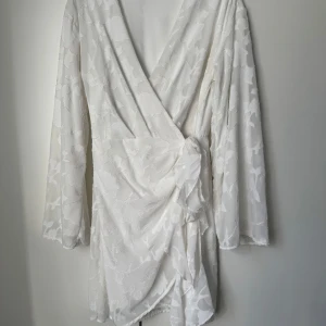 Vit omlottklänning med blommönster - Säljer en vit kort omlottklänning med subtilt blommönster. Klänningen har långa ärmar, v-ringad hals och knyt i midjan som ger en snygg siluett. Materialet är lätt och har en elegant struktur, perfekt för sommarens alla tillfällen.