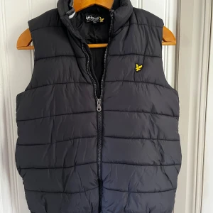 Svart dunväst från Lyle & Scott - Snygg svart dunväst från Lyle & Scott. Perfekt att slänga över en hoodie för en chill look. Junior 14/15 år