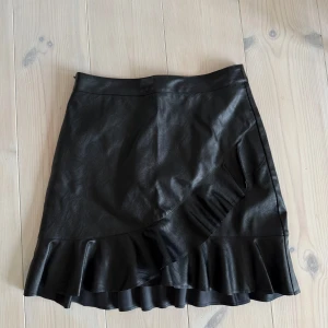 Svart kjol i skinnlook från Neo Noir - Supersnygg svart kortkjol i skinnimitation från Neo Noir. Kjolen har volangdetaljer längs nederkanten och en omlottliknande design framtill. Perfekt för dig som vill ha en edgy men feminin look. Rökfritt hem!