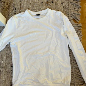 Vit långärmad tröja från BOSS - Vit långärmad tröja från BOSS med diskret logga på bröstet och BOSS-tryck längs ärmarna. Klassisk rund halsringning och mjukt bomullsmaterial som känns skönt mot huden. Perfekt för en clean och stilren look.