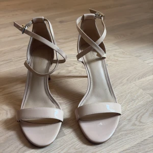 Beige klackar från Michael Kors - Snygga beige klackar från Michael Kors med smala remmar som korsas över foten och runt vristen. Skorna har öppen tå, smal klack och är tillverkade i glansigt skinn. Perfekta för dig som gillar stilrena och eleganta skor.