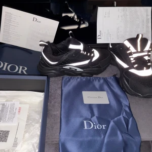 Svarta sneakers från Dior med reflexdetaljer - Snygga svarta sneakers från Dior med feta vita reflexdetaljer som lyser upp i mörkret. Skorna har chunky sula, mesh och skinnpaneler samt klassisk snörning. Perfekta för dig som vill sticka ut med exklusiv streetstyle och premiumkänsla.
