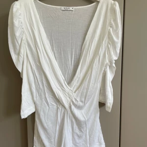 Vit topp från Vila, storlek S - Säljer en vit topp från Vila i storlek S. Toppen har djup v-ringning, korta puffärmar och är tillverkad i mjukt, tunt modaltyg. Perfekt för dig som gillar en luftig och stilren look med snygga detaljer vid axlarna.