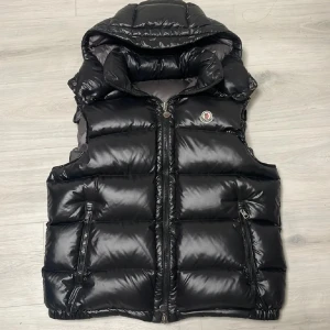 Svart dunväst från Moncler äkta - Snygg svart dunväst från Moncler med glansig finish och hög krage. Västen har två dragkedjefickor, tydlig logga på bröstet och quiltad design. Perfekt för lager-på-lager och har en sportig vibe. Stängs med dragkedja och har en klassisk Moncler-insida med serietidningsmönster. Kvittot är tyvärr slängt, behöver få sålt den snabbt så tar nog emot dom flesta buden. Dragkedja sönder därför nedsatt pris