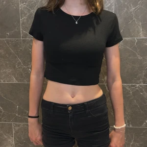 Svart croppad topp från H&M Divided - En svart, croppad topp från H&M Divided med korta ärmar och rund hals. Enkel och stilren design i mjuk bomull, perfekt till jeans eller kjol. Sitter tight och har en basic vibe som funkar till många olika looks.