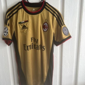 AC Milan Kaka' 22 bortatröja Adidas - AC Milan bortatröja i guld med svarta och röda detaljer, Adidas-logga och Fly Emirates-tryck. Kaka' 22 på ryggen. Kortärmad modell med klubbmärke och Champions League-patch på ärmen. Tillverkad i lätt och ventilerande material.
