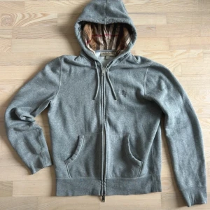 Grå hoodie från Burberry med dragkedja - Snygg grå hoodie från Burberry med klassiskt rutigt foder i huvan. Tröjan har dragkedja framtill, snörning vid huvan och två fickor. Tillverkad i mjuk bomull och polyesterblandning. Perfekt för dig som gillar stilrena och exklusiva plagg.