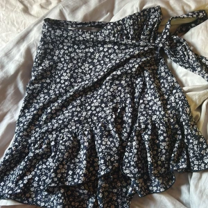 Blommig omlottkjol från ONLY - Supersöt kort omlottkjol från ONLY i marinblått med vitt blommönster. Kjolen har volangkant och knyts i midjan för en snygg passform. Perfekt för dig som gillar en feminin och trendig stil.