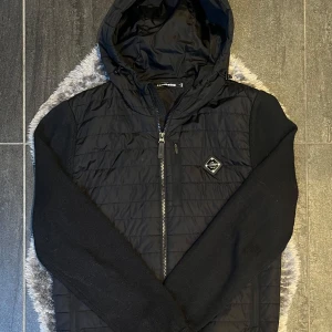 J.Lindeberg Svart Hybrid Cardigan Jacka  - Stilren hybridjacka/cardigan från J.Lindeberg i färgen svart, storlek S. Perfekt som lätt jacka eller mellanlager. I mycket fint skick – endast några få tecken på användning, inget som stör helhetsintrycket.