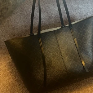 Stor svart shoppingväska i skinn - Säljer en stor och rymlig svart shoppingväska med diskret rutmönster. Väskan har långa, smala axelband och är tillverkad i ett tåligt skinnmaterial. Perfekt för dig som vill ha plats för allt du behöver under dagen.