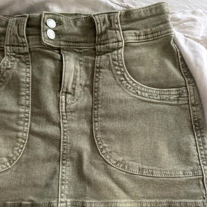 Olivgrön jeanskjol från Bershka - Olivgrön kort mini jeanskjol från Bershka med stora fickor fram och bak, bred linning med dubbla knappar och bälteshällor. 