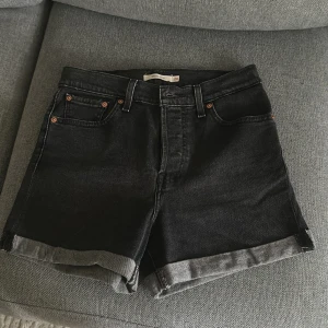 Svarta jeansshorts från Levi's - Svarta jeansshorts från Levi's med klassisk femficksmodell, uppvikta benslut och knäppning med fyra silverfärgade knappar. Snyggt läderpatch bak och ikonisk röd Levi's-tagg på bakfickan. Perfekta för en chill sommarstil.