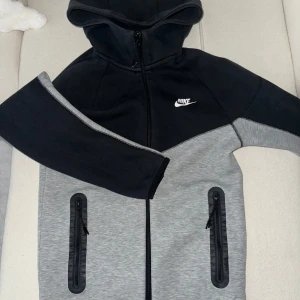 Nike hoodiejacka svart/grå XS - Nike hoodiejacka i svart och grått med dragkedja framtill och två fickor med dragkedja. Jackan har huva och sportig look, perfekt för chill dagar eller när du är på språng. Snygg kontrast mellan färgerna och klassisk Nike-logga på bröstet. XS passar S. 