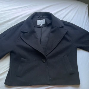 Svart kappa från Vila med knappar - Stilren svart kappa från Vila med klassisk krage och en knapp framtill. Jackan har raka fickor och är i ett mjukt ull-liknande material. Perfekt för dig som gillar en clean och tidlös look. Passar till allt och är enkel att styla.