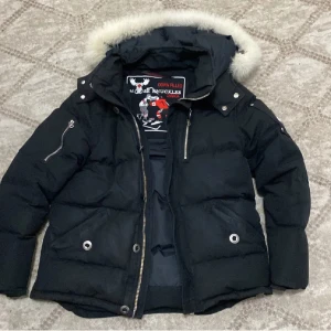 Svart dunjacka Moose Knuckles päls - Säljer en svart dunjacka från Moose Knuckles med fluffig päls på huvan och snygga silverdetaljer. Jackan har dragkedjor på ärmar och fram, samt broderad hockeypatch på insidan. Perfekt för kalla dagar och riktigt street. Ikonisk logga på ärmen.