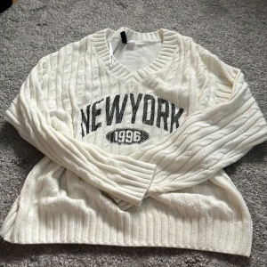 Vit stickad tröja med New York-tryck - Vit kabelstickad tröja med svart tryck 'NEW YORK 1996' på bröstet. Tröjan har v-ringad hals, långa ärmar och breda ribbade muddar. Ganska använd men visar inga täcken på dålig slitningar:) säljs ej längre på HM