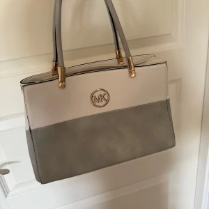 handväska Michael Kors - Snygg handväska Michael Kors liknande i vitt och grått med guldfärgade detaljer och MK-logga framtill. Väskan har flera fack, dragkedja på baksidan och rejäla handtag. Perfekt för dig som vill ha en stilren och rymlig väska till vardagen.