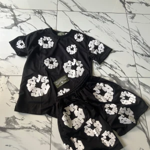 Svarta shorts från Denim Tears M - Svarta shorts från Denim Tears med unika vita blommiga tryck över hela plagget. De har elastisk midja med snörning för skön passform. Perfekta för dig som vill sticka ut med en cool och modern stil. Matcha med t-shirten för en komplett look.
