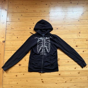 Svart hoodie med skelettmotiv och huva - Svart hoodie med dragkedja och huva, HEMMAGJORD  med glittrigt skelettmotiv i silverfärgade paljetter på framsidan. Långärmad modell med fickor framtill. Perfekt för dig som vill sticka ut med en edgy och cool stil.