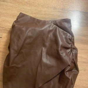 Brun omlottkjol i skinnimitation - Snygg brun kortkjol i skinnimitation med omlott-design och rynkade detaljer på sidan. Kjolen har en cool och trendig look som passar perfekt till boots eller sneakers. Perfekt för dig som vill sticka ut lite extra.