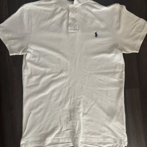 Vit piké från Polo Ralph Lauren - Storlek M. Vit pikétröja från Polo Ralph Lauren med klassisk krage, två knappar och den ikoniska broderade loggan i mörkblått på bröstet. Slimmad passform och tillverkad i bomull för en fräsch och stilren look. Nypris (1595kr) 