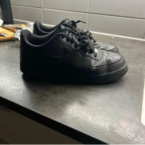 Nike Air Force 1  - Klassiska helsvarta Nike Air Force 1 sneakers i skinn med låg profil och rund tå. Skorna har snörning och en platt sula, perfekt för en clean och stilren look. Ikonisk modell som funkar till det mesta och ger en tidlös vibe.