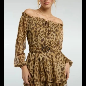 Leopardmönstrad offshoulder klänning - Snygg kort klänning med leopardmönster i beige och bruna toner. Offshoulder-modell med långa ärmar, volangdetaljer och markerad midja med bälte. Perfekt för dig som vill sticka ut med en trendig look. Klänningen har shorts! Med prislapp