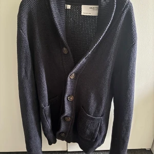 Mörkblå kofta från Selected Homme - Säljer en mörkblå stickad kofta från Selected Homme i storlek M. Koftan har grovstickad struktur, djup V-ringning, stora knappar framtill och två fickor. Perfekt för lager-på-lager och chill stil.