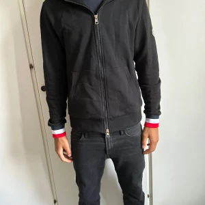 Svart zip-hoodie från Moncler - Svart hoodie från Moncler med hel dragkedja, fickor framtill och Moncler-logga på ärmen. Muddarna har snygga ränder i rött, vitt och blått för en extra touch. Tillverkad i mjuk bomull, perfekt för en chill och stilren look. M som passar som s