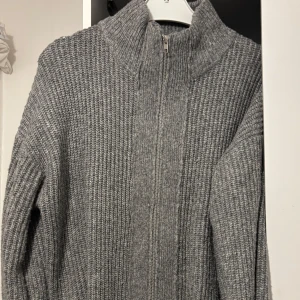 Grå stickad kofta med dragkedja - Mysig grå stickad kofta med hög krage och hel dragkedja framtill. Tröjan har en ribbad struktur och långa ärmar, perfekt för kyliga dagar. Enkel att matcha och superbekväm att bära.