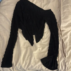 Svart oneshoulder topp med volang - Säljer en svart oneshoulder topp med lång ärm och volangdetalj vid ärmslutet. Toppen har en asymmetrisk skärning och är tillverkad i ett lätt, vågigt tyg som ger en cool struktur. Perfekt för dig som vill sticka ut med en unik siluett.