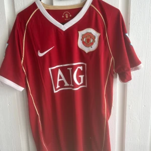 Manchester United Ronaldo 7 Nike tröja - Klassisk Manchester United fotbollströja med Ronaldo 7 på ryggen. Röd med vita detaljer och guldiga linjer, officiell AIG sponsorlogga på bröstet och klubbmärke. Tillverkad i lätt och ventilerande material, perfekt för match eller träning.