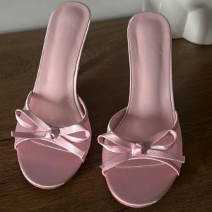 Rosa satinpumps med rosett - Snygga ljusrosa pumps i satin med öppen tå och söt rosettdetalj framtill. Skorna har hög klack och en elegant, feminin look som verkligen sticker ut. Perfekta för dig som vill ha en chic och trendig stil.