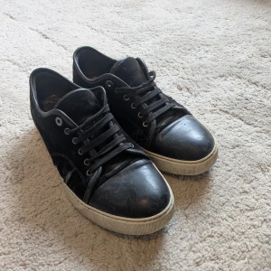 Svarta sneakers från Lanvin i mocka och skinn - Svarta sneakers från Lanvin med ovandel i mocka och tåparti i skinn. Klassisk låg modell med snörning och vit sula. Snygg kontrast mellan material och en stilren look som funkar till det mesta. Märkeslogga på sulan. Skorna är slitna och har många slit som ni ser på bilderna men detta kan färgas över med mocka spray för att få det är se fräschare ut.