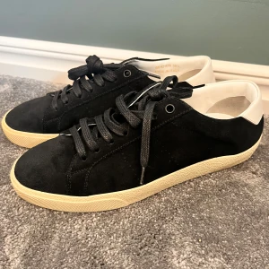 Svarta Saint Laurent sneakers i mocka - Snygga svarta sneakers från Saint Laurent i mjuk mocka med vita skinn-detaljer på hälen och beige sula. Klassisk låg modell med rund tå och svarta snören. Perfekta för dig som gillar stilrena och exklusiva skor med en lyxig känsla.