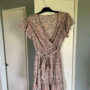 Blommig sommarklänning  - Supersöt omlottklänning med blommigt mönster i ljusa färger som aldrig kommit till användning. Klänningen har volangärmar, knytband i midjan och v-ringning. Perfekt för sommarens alla tillfällen. Materialet känns lätt och luftigt.