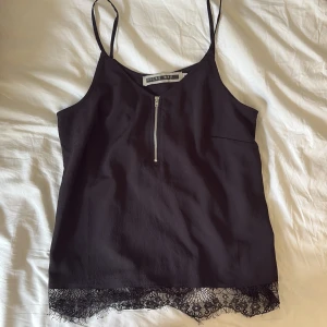 Svart linnetopp med spets från Noisy May - Svart linnetopp från Noisy May med tunna axelband, dragkedja framtill och snygg spetskant längst ner. Toppen har en enkel, stilren design och är perfekt för att lyfta vilken outfit som helst.