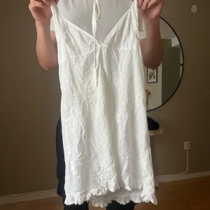 Vit kort sommarklänning med spets - Söt vit kort klänning med tunna axelband och broderade spetsdetaljer. Klänningen har v-ringning, knyt i nacken och volangkant nertill. Perfekt för varma dagar och har en romantisk vibe.