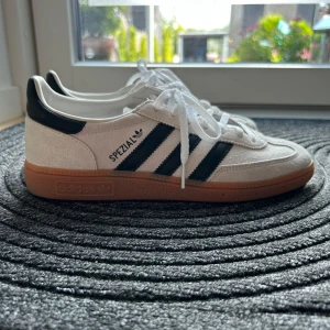 Adidas Spezial beige - Adidas Spezial sneakers i vitt mocka med svarta klassiska ränder och svart häl. Snygg gummisula i brunt och vita snören. Ikonisk retrostil med låg profil och rund tå, perfekt för dig som gillar streetwear och klassiska vibes. Nypris: 1300, har använt de 1 gång.