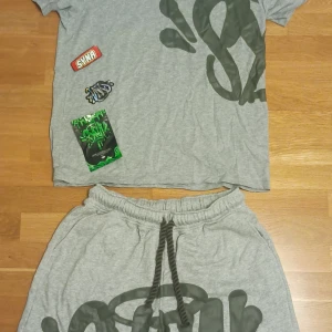 Grå set syna world M - Grå set från syna worldi mjuk bomull med bred resår och snörning i midjan. Stora mörkgrå grafitti-liknande tryck framtill ger en streetig vibe. Perfekta för chill dagar eller när du vill ha en avslappnad look.