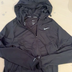 Nike Miller - Svart vindjacka från Nike med huva och dragkedja framtill. Jackan har reflekterande detaljer på bröstet och ärmarna, samt diskret Nike-logga. Lätt och smidig modell i syntetmaterial, perfekt för löpning eller utomhusaktiviteter.